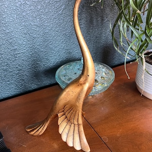 May include: A gold-colored bird figurine with a long, slender neck and curved body. The sculpture features detailed wing feathers and a smooth, metallic finish with subtle dark distressing, standing in an upright, alert posture.