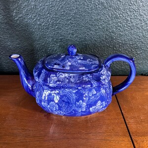 May include: A blue ceramic teapot with a floral pattern. The teapot has a curved handle, a spout, and a lid with a small knob. The floral design is white against the blue background. The teapot is sitting on a wooden surface.