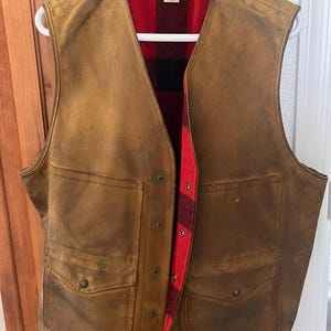 Puede incluir: Un chaleco marr&oacute;n con forro de cuadros rojos y negros. El chaleco tiene dos bolsillos delanteros y est&aacute; abotonado. El chaleco est&aacute; colgado de una percha blanca.