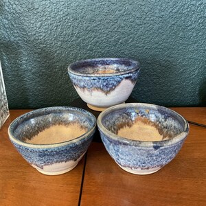 May include: Three handmade ceramic bowls. The bowls have a blue and white glaze with a tan interior. The bowls are of varying sizes and are arranged on a wooden surface. The bowls are likely for serving food.