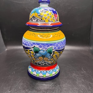 May include: A colorful, hand-painted ceramic jar with a lid. The jar features a vibrant design with yellow, blue, red, and green accents. The base and lid are adorned with intricate floral patterns, making it a decorative piece.
