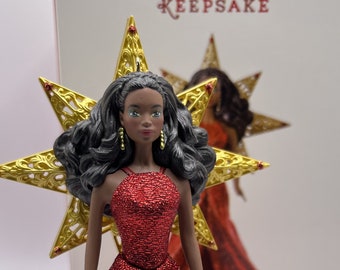 May include: A black doll with long black hair wearing a red sequined dress and a gold star ornament. The doll is a Christmas tree topper.