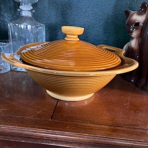 May include: A vintage, lidded serving bowl in shades of brown and tan. The bowl has a round shape with a lid and handles. The lid and bowl feature a ribbed design. The bowl is sitting on a wooden surface.