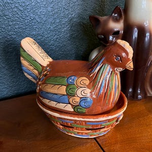 May include: A ceramic hen-shaped container, primarily brown with colorful accents. The hen sits atop a matching basket-like base, with details in blue, green, and orange. The decorative piece is likely for kitchen or home decor.