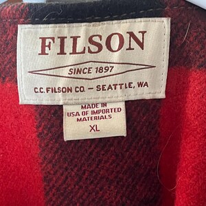Puede incluir: Primer plano de una chaqueta a cuadros rojos y negros con una etiqueta que dice "FILSON SINCE 1897" y "MADE IN USA OF IMPORTED MATERIALS XL". La chaqueta est&aacute; hecha de un material grueso y c&aacute;lido.