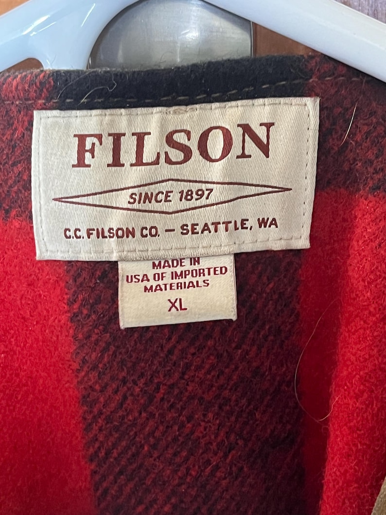 Puede incluir: Primer plano de una chaqueta a cuadros rojos y negros con una etiqueta que dice "FILSON SINCE 1897" y "MADE IN USA OF IMPORTED MATERIALS XL". La chaqueta est&aacute; hecha de un material grueso y c&aacute;lido.