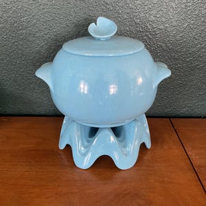 May include: A light blue ceramic fondue pot with a matching lid and a decorative stand. The pot has two handles and a flower-shaped knob on the lid. The stand has a wavy design.
