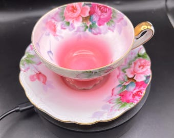May include: A pink teacup and saucer set with a floral rose design. The teacup has a gold handle and rim. The saucer has a scalloped edge and a matching rose pattern. The set is on a black surface.