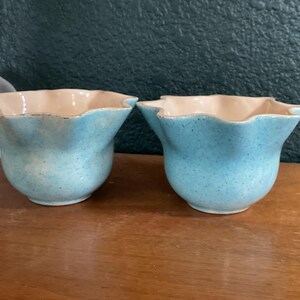 May include: Two matching ceramic bowls with a light blue speckled exterior and a smooth cream-colored interior. Each bowl features a distinct scalloped, wavy rim design and a rounded base, presenting a consistent aesthetic across both pieces.