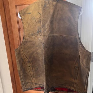 Puede incluir: Un chaleco de cuero marr&oacute;n con un acabado envejecido, colgado de una percha blanca. El chaleco tiene un forro a cuadros rojo y negro visible en la parte inferior. Dos botones color lat&oacute;n est&aacute;n en la parte delantera.