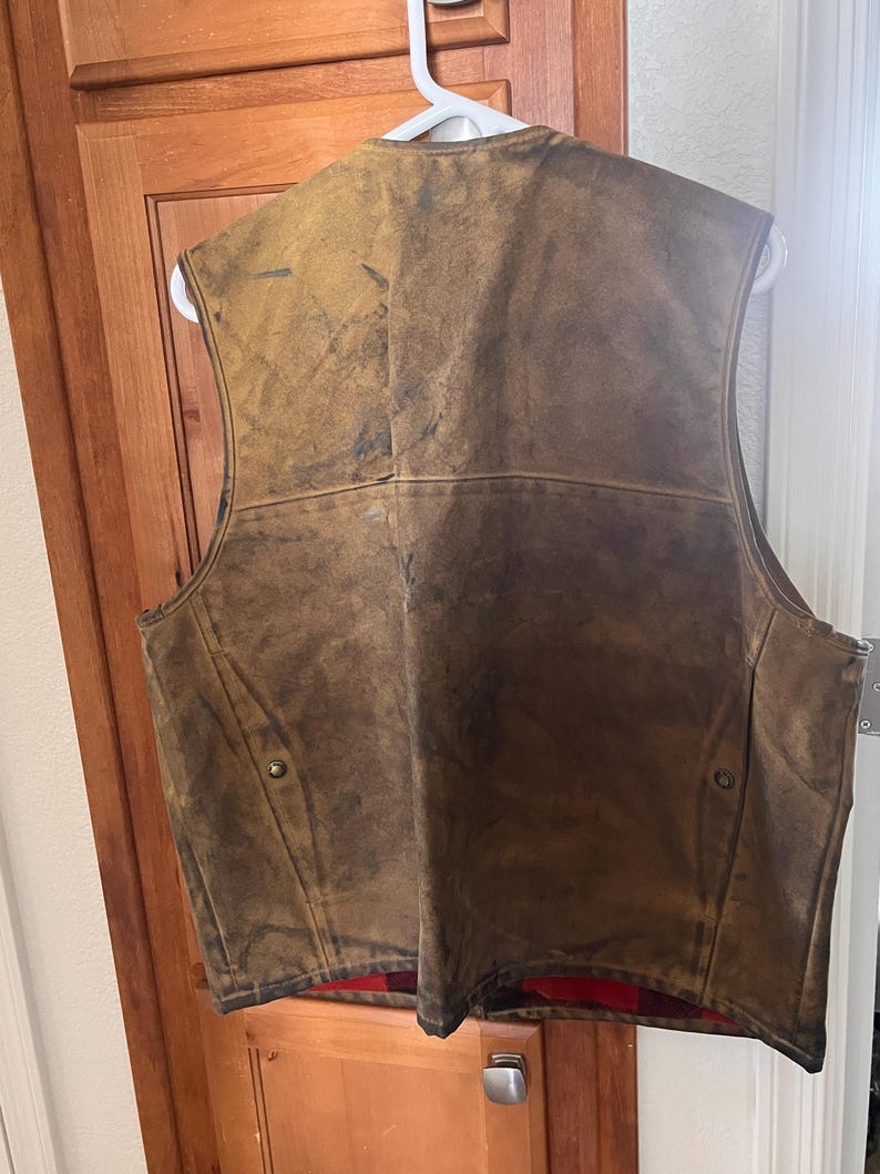 Puede incluir: Un chaleco de cuero marr&oacute;n con un acabado envejecido, colgado de una percha blanca. El chaleco tiene un forro a cuadros rojo y negro visible en la parte inferior. Dos botones color lat&oacute;n est&aacute;n en la parte delantera.