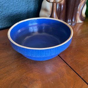 May include: A round, cobalt blue bowl with a textured exterior and a cream-colored rim. The bowl is made of ceramic and sits on a wooden surface. A decorative cat figurine is in the background.