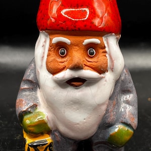 May include: A ceramic gnome figurine with a red hat, white beard, and a lantern in its hand. The gnome is wearing a blue and green outfit.
