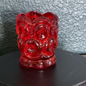 May include: A vibrant red glass votive holder featuring a repeating moon and star pattern embossed across its cylindrical surface. The glass has a scalloped top rim and a flared base, with a translucent finish that catches light through the textured design.