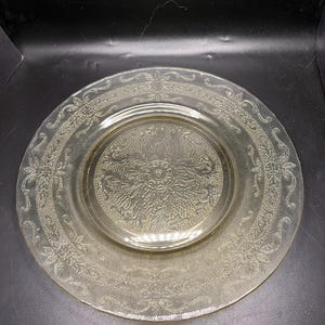 May include: A clear, vintage glass plate with an embossed floral design. The plate features a detailed pattern around the rim and a central floral motif. The plate has a slightly yellow tint and is likely made of pressed glass.