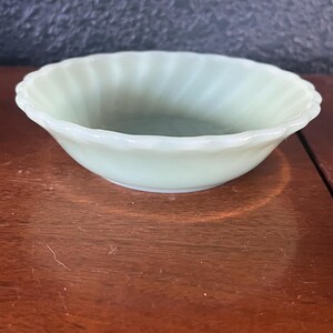 May include: A small, light green glass bowl with a scalloped rim. The bowl has a rounded shape and a smooth interior. The glass has a slight translucence, and the bowl sits on a wooden surface.