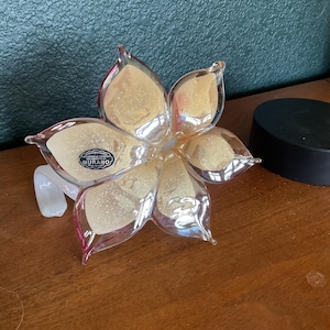 May include: A Murano glass flower sculpture with six petals. The petals are a translucent yellow with pink edges and glitter accents. A small, black and white label is visible on the flower. The sculpture is on a wooden surface.