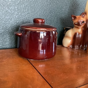 May include: A glossy, dark brown ceramic casserole dish with a matching lid. The dish has two small handles and a rounded shape. The lid features a small knob for easy handling. The casserole dish is sitting on a wooden surface.