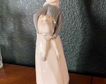 May include: A porcelain figurine of a woman holding a lamb. The woman wears a white headscarf, a gray jacket, and a long white dress. The lamb is light beige. The figurine is on a wooden surface.