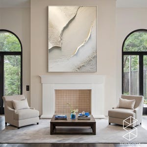 May include: A white and gold abstract painting with a textured surface hangs above a fireplace in a living room. The painting is framed in a thin, white frame. Two beige armchairs are placed on either side of a coffee table in front of the fireplace.