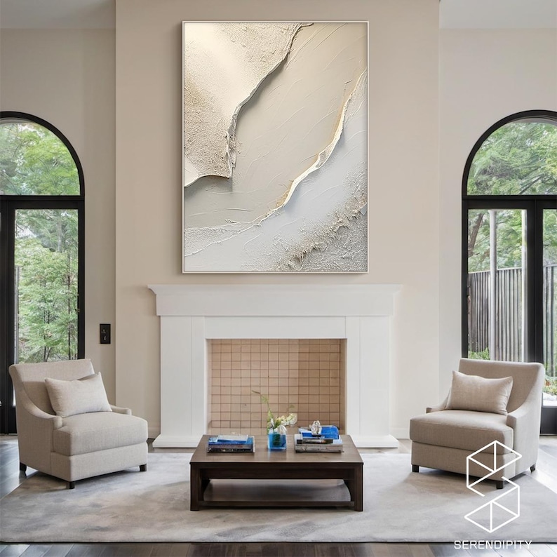May include: A white and gold abstract painting with a textured surface hangs above a fireplace in a living room. The painting is framed in a thin, white frame. Two beige armchairs are placed on either side of a coffee table in front of the fireplace.