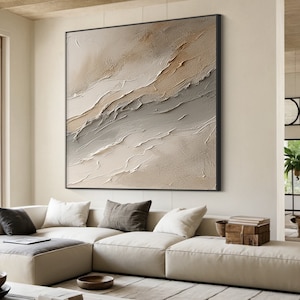 May include: Abstract painting with a textured, layered look in shades of beige, brown, and gray. The painting is framed in black and hangs on a wall above a white sofa.