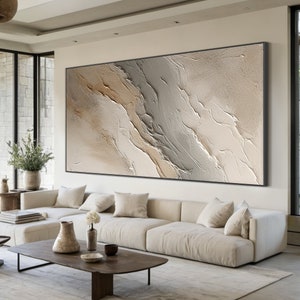 May include: Abstract painting with a textured, layered design in shades of beige, brown, and gray. The painting is framed in a black frame and hangs on a white wall in a modern living room.