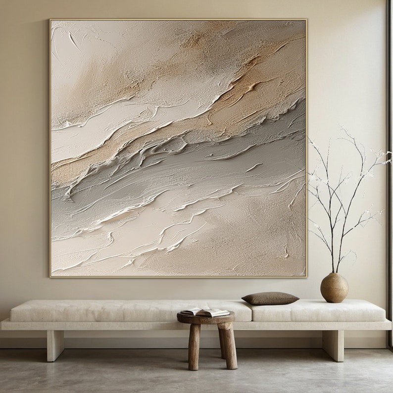 May include: Abstract painting with a textured, layered design in shades of beige, brown, and white. The painting is framed in a thin gold frame and hangs on a white wall.