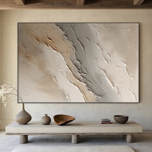 May include: Abstract painting with a textured, layered design in shades of beige, brown, and gray. The painting is framed in a black frame and hangs on a wall above a wooden table.