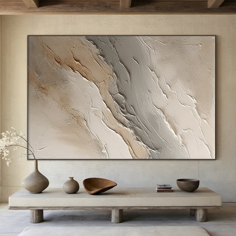 May include: Abstract painting with a textured, layered design in shades of beige, brown, and gray. The painting is framed in a black frame and hangs on a wall above a wooden table.