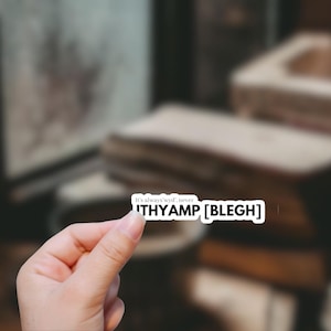 May include: A white sticker with the text "It's always wyd, never ITHYAMP [BLEGH]" in black. The sticker is held in a hand against a blurred background of books.