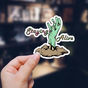 May include: A sticker with a green zombie hand reaching out of the ground. The text "Staying Alive" is on the left side of the sticker and "Alive" is on the right side.