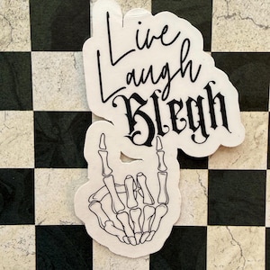 May include: White sticker with black text that reads "Live Laugh Blegh" in a mix of script and gothic fonts. Below the text is a skeleton hand making a rock and roll gesture. The sticker is on a black and white checkered background.