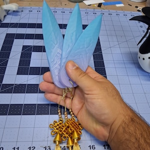 Puede incluir: Tres adornos en forma de pluma azul y morada con borlas doradas y nudos decorativos. Las plumas se sujetan con hilo dorado. El fondo es una esterilla de corte azul.