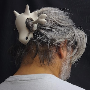 May include: A white, stylized skull hair clip with a bone accent, worn in gray hair. The clip is a unique accessory, likely for cosplay or themed events. The skull has a large eye socket and a bone protruding from the side.