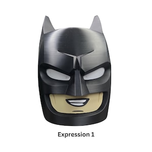 May include: A black Batman mask with white mesh eye openings and a tan-colored mouth area. The mask has pointed ears and a textured surface. The text "Expression 1" is printed at the bottom.