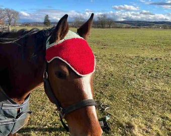 May include: A brown horse wearing a red and white crocheted fly bonnet. The bonnet covers the horse's ears and forehead, with a white trim. The horse is in a grassy field on a sunny day.