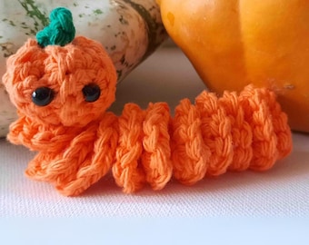 Op de afbeelding: Een oranje gebreide pompoen met een groene steel en zwarte ogen. De pompoen is bevestigd aan een lange oranje gebreide staart.