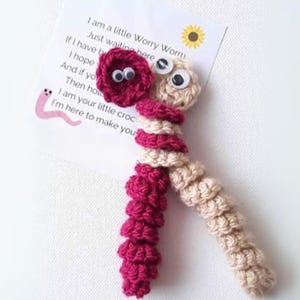 Può includere: Un giocattolo a forma di verme crochetato con occhietti mobili. Il verme è fatto di due colori di filato, rosa e beige. Il verme tiene un biglietto che dice "Sono un piccolo verme che si preoccupa. Aspetto solo qui. Se ho... Spero... E se tu... Allora tieni... Sono il tuo piccolo coccodrillo. Sono qui per farti..."