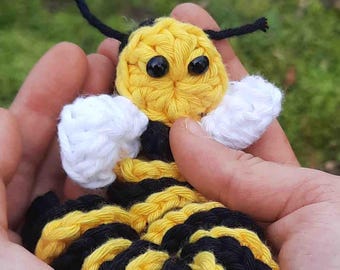 May include: A crocheted bee toy with black and yellow stripes, white wings, and black eyes. The bee is held in a person's hand.