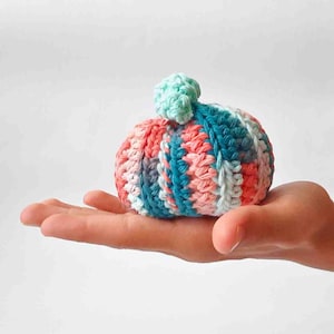 May include: A small, colorful crocheted hat with a pom-pom. The hat features stripes of coral, teal, light blue, and white yarn. The pom-pom is a light green color. The hat is held in the palm of a hand.