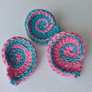 May include: Three handmade crochet spiral shapes in pink and teal. The spiral design is a contrasting teal color, with a pink base. The crochet work is detailed and the shapes are arranged on a white surface.