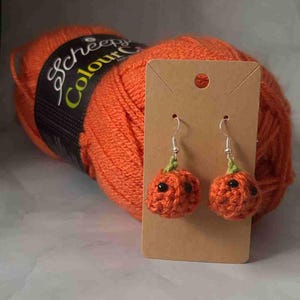 May include: Two orange crochet pumpkin earrings with black bead eyes and green stems, suspended from silver hooks. The earrings are presented on a brown card, with a large ball of orange yarn behind them.