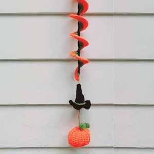 Könnte beinhalten: Eine gehäkelte Halloween-Girlande mit orangefarbenen und grünen Kürbissen und einem schwarzen Hexenhut, die an einer weißen Wand hängt.