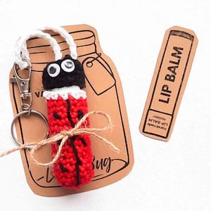 Puede incluir: Un porta bálsamo labial de ganchillo de color rojo y negro con forma de mariquita con ojos saltones y una cadena de llaves. El porta bálsamo está atado con un cordel y se muestra en una etiqueta de papel marrón con el texto "Lip Bug" y un dibujo de un frasco. Una etiqueta de papel marrón separada dice "Handmade Lip Balm Holder Lip Balm".
