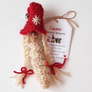May include: A crocheted gnome worry worm with a red hat and white snowflake. The gnome has long blonde hair and is holding a tag that says "Gnome Worry Worm" and "Your Magical gnome, just for you. If you're feeling sad, hold me tight and I'll see you through."