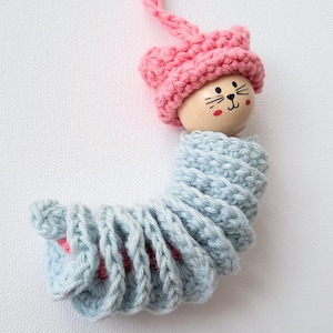 May include: A crocheted caterpillar toy with a pink crocheted hat and a wooden head with a painted cat face. The caterpillar is curled up in a spiral shape.