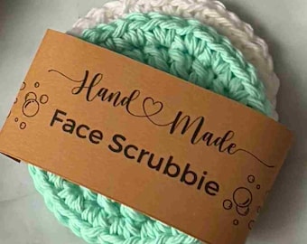 Könnte beinhalten: Ein Stapel von drei gehäkelten Gesichtsschwämmen in Weiß, Mintgrün und Weiß. Ein braunes Etikett mit den Worten "Hand Made Face Scrubbie" liegt obenauf. Die Schwämme sind rund und aus dickem Garn gefertigt.