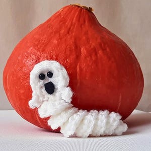 Könnte beinhalten: Ein weißer gehäkelter Geist mit schwarzen Augen und einem Mund ist um einen leuchtend orangefarbenen Kürbis gewickelt.