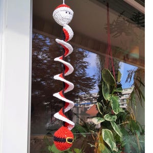May include: A crocheted Santa Claus ornament with a red and white spiral body and a black belt. The ornament is hanging from a window.